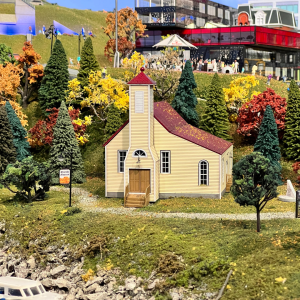 An image of a miniature building with a red roof and brown door. There are trees and green foliage around the structure.