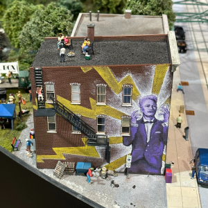 An image of a miniature building with a mural painted on the side of it. There are miniature figures standing on the roof, and there are people behind the people as well.