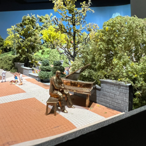 An image of a miniature brass figure next to a piano. There is green foliage around the figure and in the distance.