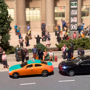 An image of a single orange car with a black car behind it. There are people standing in the distance.