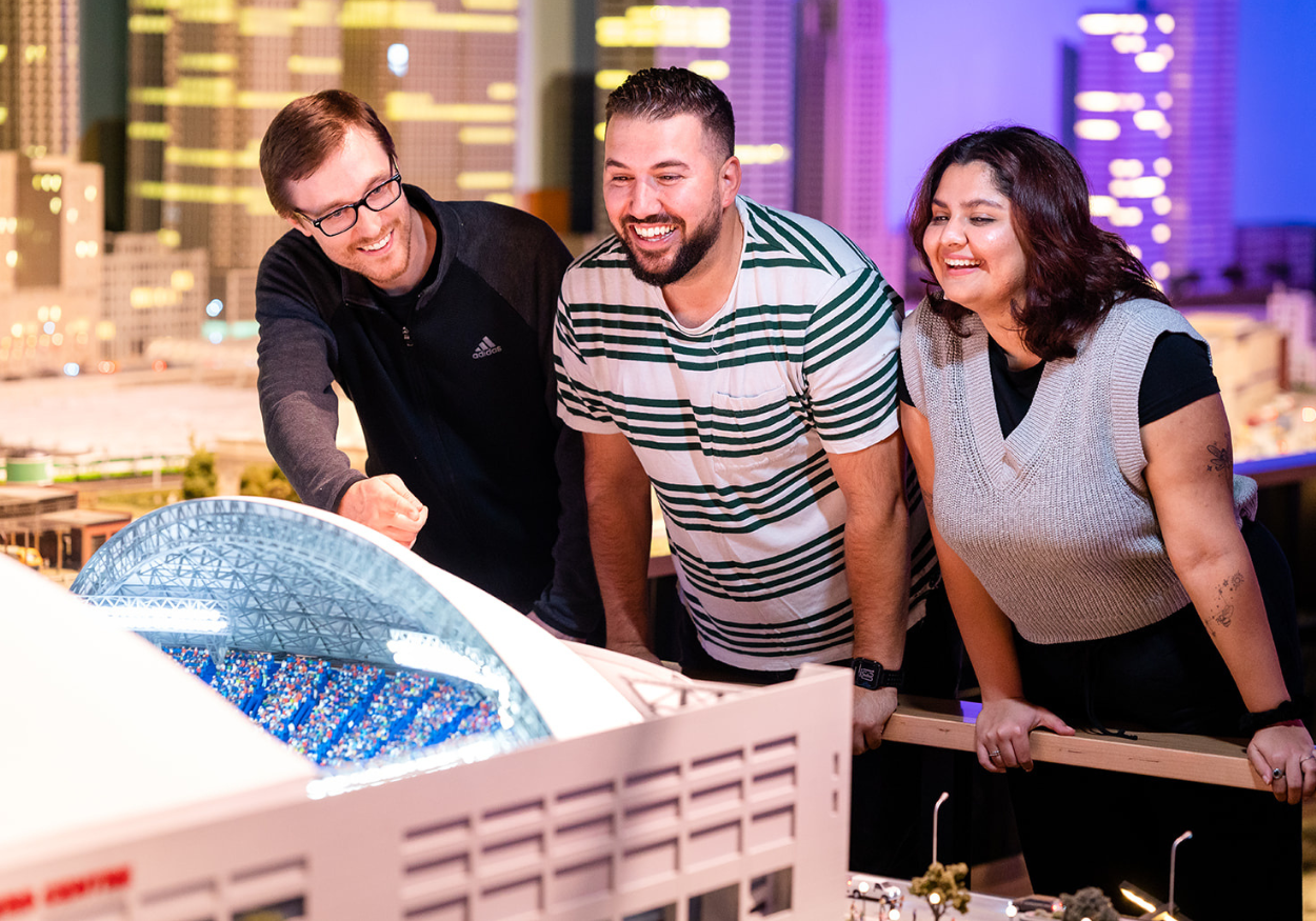 Three people looking closely into a miniature structure of a stadium.