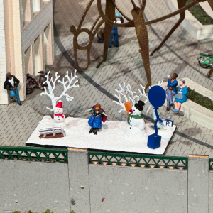 A miniature scene of a young person standing on snow in between two snowmen.