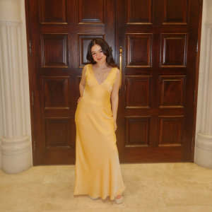 A young girl with dark hair wearing a long yellow gown standing in front of a wooden door.