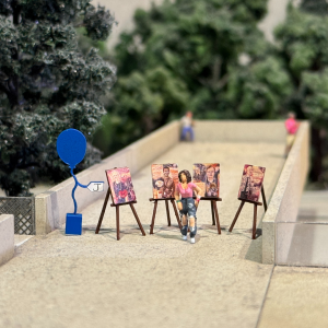 A miniature display of a young person standing alongside their artwork with a little blue balloon positioned to the left.